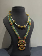 An ornate traditional Indian jewelry set displayed on a black mannequin bust, featuring a long gold-plated necklace with green and white beaded strands, red and green enamel details, and a large pendant, paired with a shorter matching pearl and gold necklace.