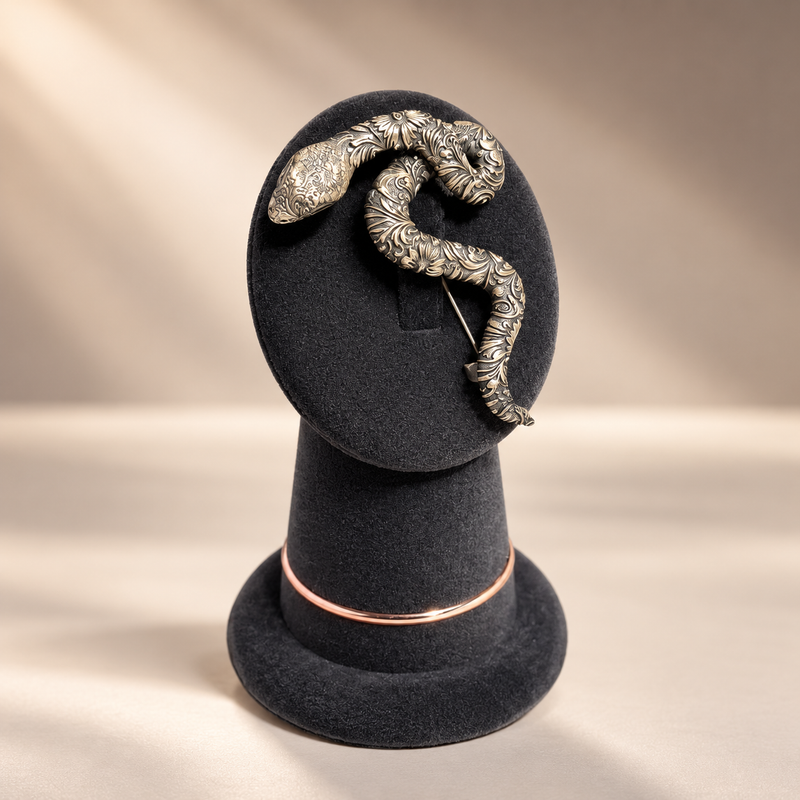 An ornate silver snake brooch with detailed floral engraving, displayed on a black velvet jewelry stand against a warm beige background.