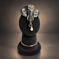 An elephant-shaped brooch with a silver-toned body, pink and green gemstone eyes, ivory tusks, and a dangling white pearl, displayed on a black velvet jewelry stand against a dark gradient background.