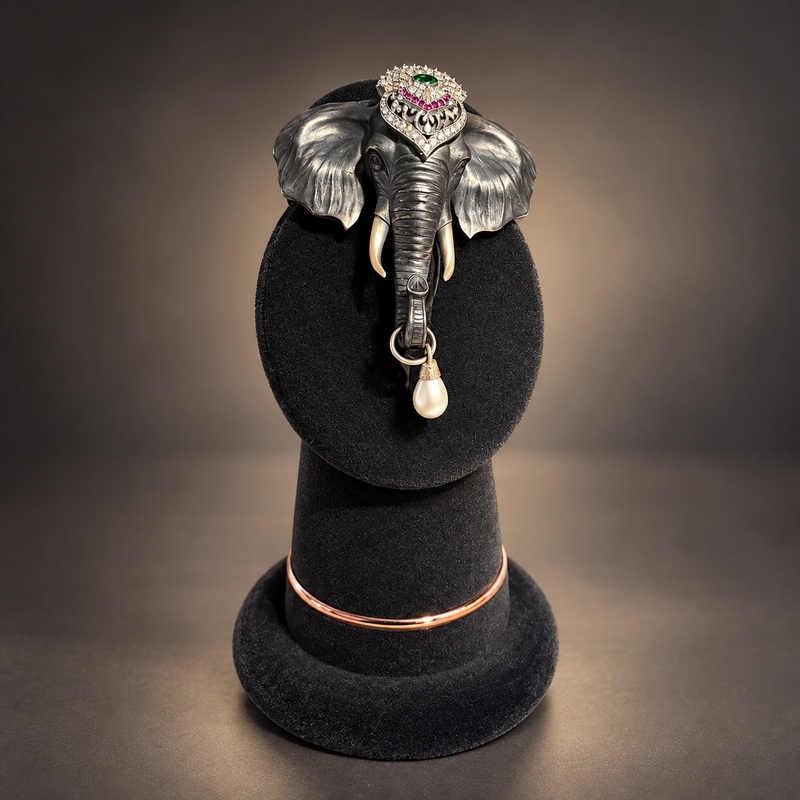 An elephant-shaped brooch with a silver-toned body, pink and green gemstone eyes, ivory tusks, and a dangling white pearl, displayed on a black velvet jewelry stand against a dark gradient background.
