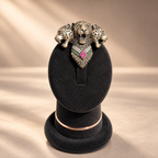A three-headed leopard pendant with a heart-shaped center featuring pink and green stones, displayed on a black velvet jewelry stand against a warm beige background.