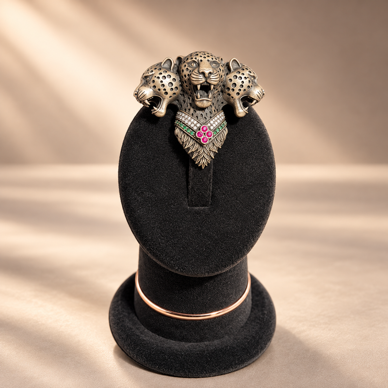 A three-headed leopard pendant with a heart-shaped center featuring pink and green stones, displayed on a black velvet jewelry stand against a warm beige background.