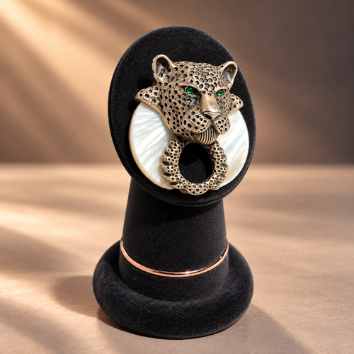 A leopard head ring holder brooch with green eyes, featuring a textured gold-toned leopard head, a white mother-of-pearl ring, and a rose gold band, displayed on a black velvet stand.